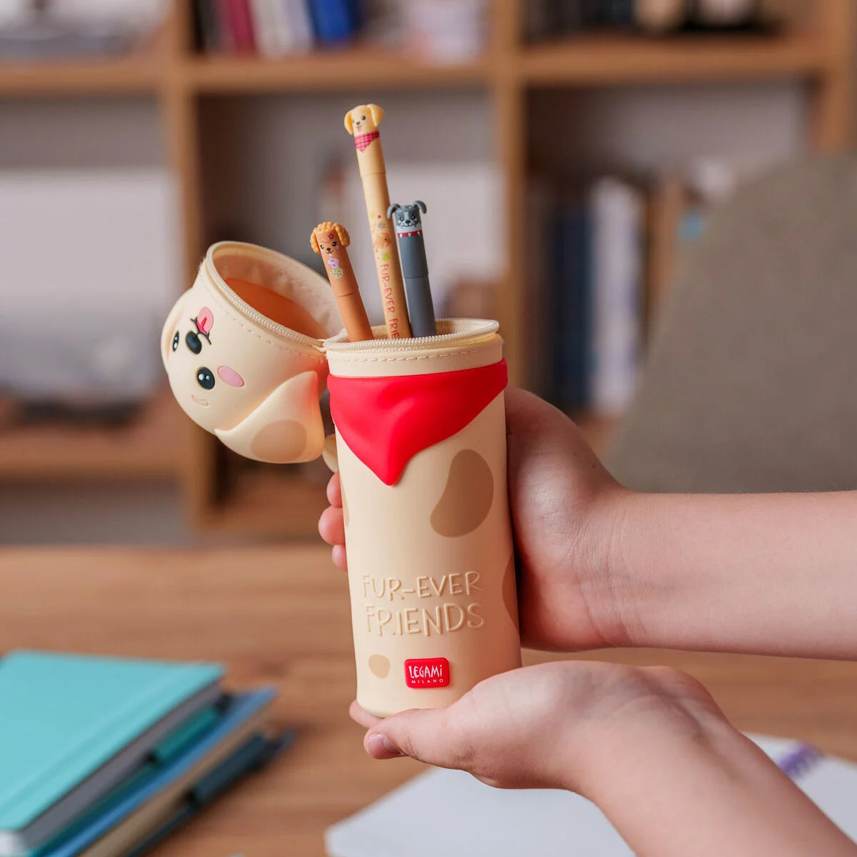 Kawaii Federmäppchen Golden Hunde-Motiv Fur-Ever Friends von Legami limitiert im Einsatz zwei Haende halten geoeffnetes beige-cremefarbenes Federmäppchen mit rotem Innenteil und Stiften auf Schreibtisch-Hintergrund mit Buecherregal