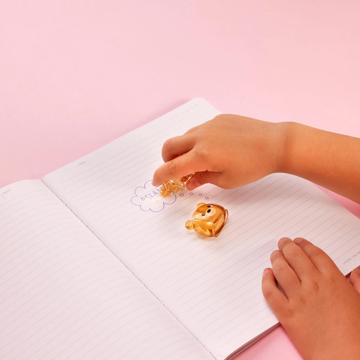 Hand benutzt den Korrekturroller Bear Sweet Bear von Legami auf einem aufgeschlagenen Notizbuch mit pinkem Hintergrund