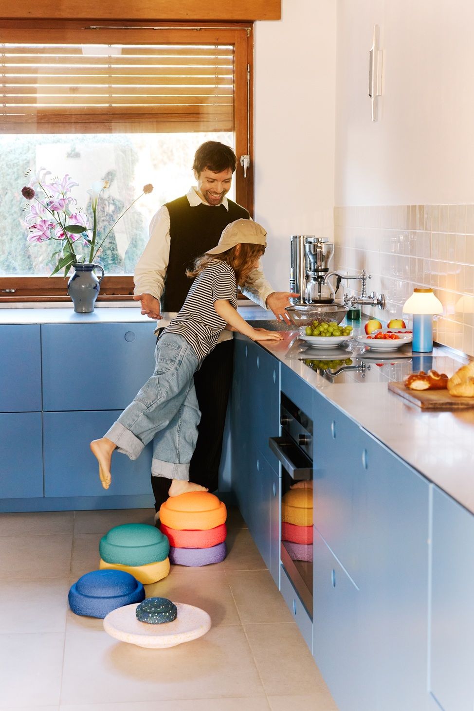 Vater und Kind in der Küche mit Stapelstein® "Spring Special" Rainbow Set classic - Stapelsteine in Regenbogenfarben mit Balanceboard und Mini-Stapelstein, Motorikspielzeug zur Förderung von Gleichgewicht, Koordination und freiem Spiel im Familienalltag