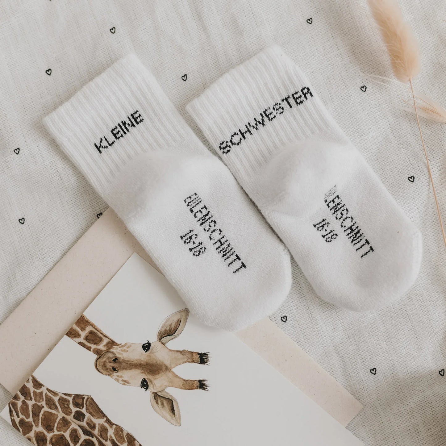 Eulenschnitt Baby-Socken „Kleine Schwester" in Weiß, Größe 16-18, auf herzgemustertem Stoff mit Giraffen-Karte bei Burgfrollein