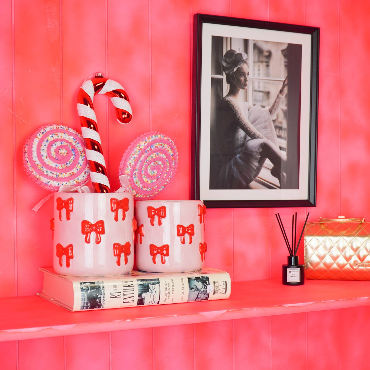Zwei rosa Blumentöpfe mit rotem Schleifen-Motiv von House Vitamin, gefüllt mit Lollis und einer Zuckerstange, auf einem Bücherstapel vor pinkem Hintergrund in Szene gesetzt – verspielter Hingucker als Wohnungsdeko und Geschenkidee