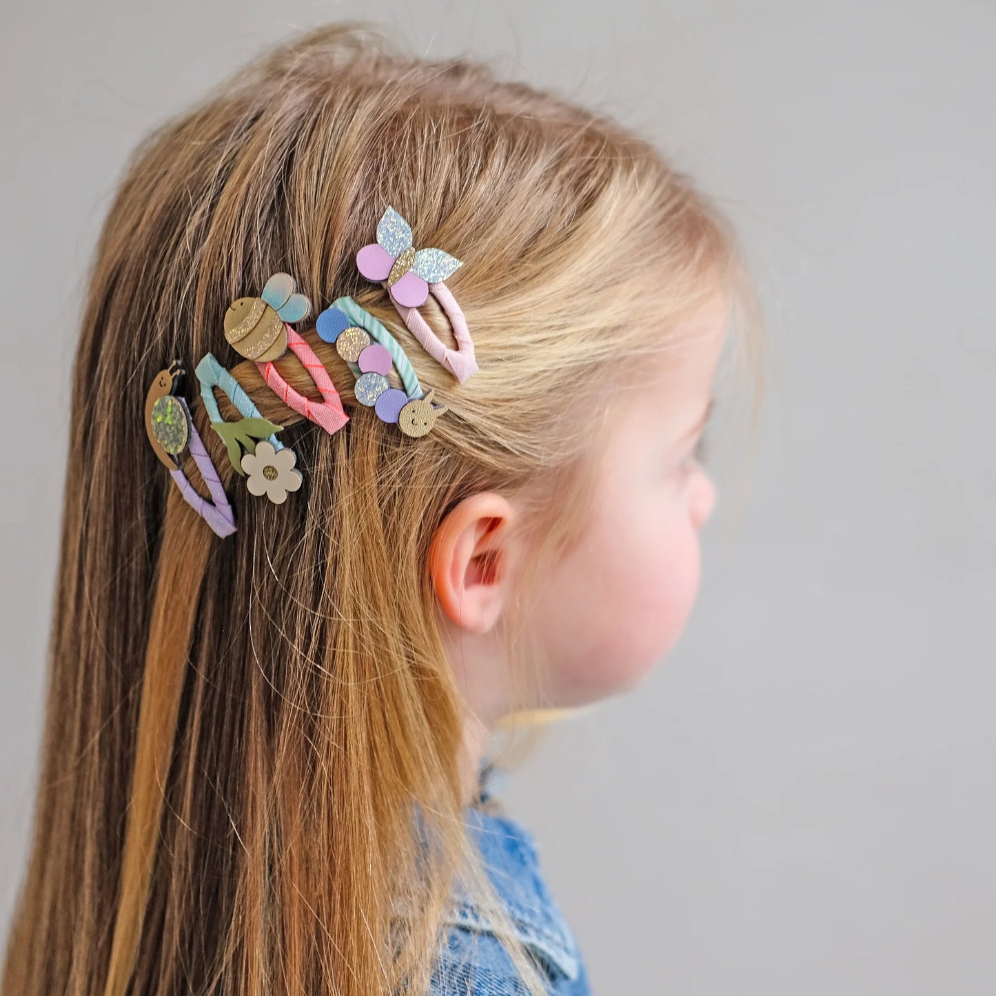Haarspangen mit Frühlings-Motiven von Mimi & Lula, 5er Set mit Biene, Blume, Schmetterling, Raupe und Schnecke in Pastelltönen mit Glitzer-Details, getragen im Haar eines Kindes – kinderfreundlicher Haarschmuck und liebevolle Geschenkidee
