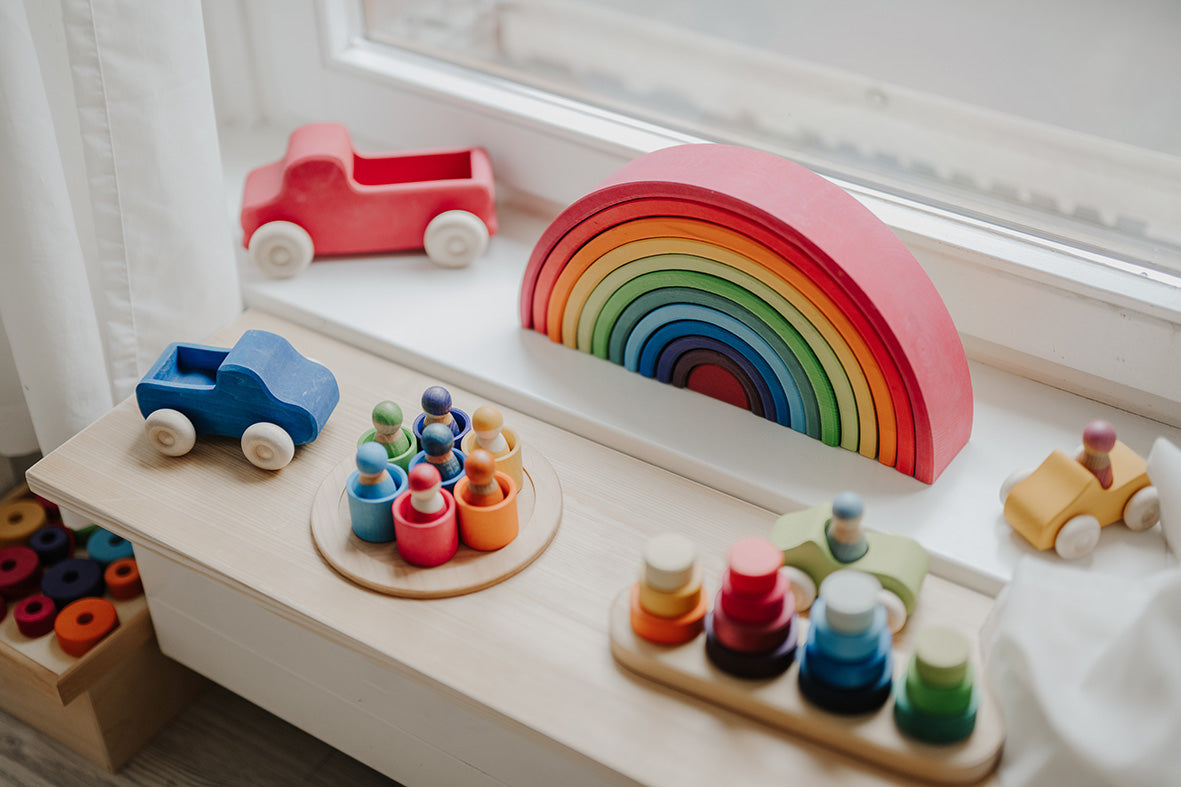 Grimms Spielzeug im Alltag: roter und blauer Lastwagen, großer Regenbogen, Regenbogenmännchen im Schälchen und Steckspiel auf der Fensterbank