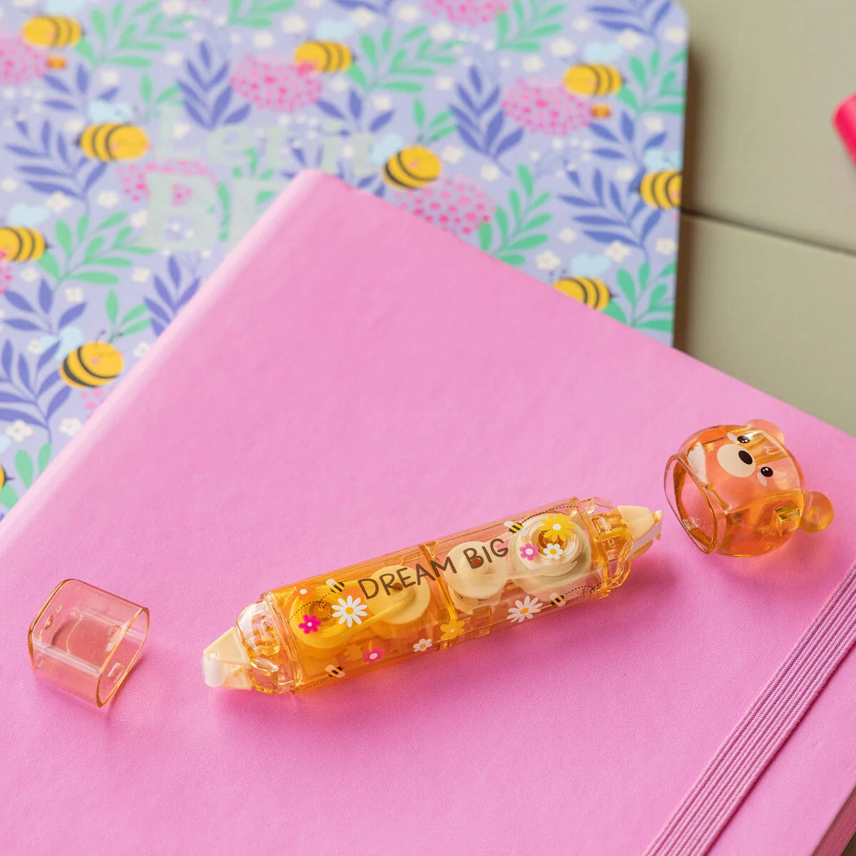Zwei-in-Eins Korrektur- und Kleberoller Bear Sweet Bear von Legami auf einem pinken Notizbuch mit buntem Bienenmuster-Papier im Hintergrund