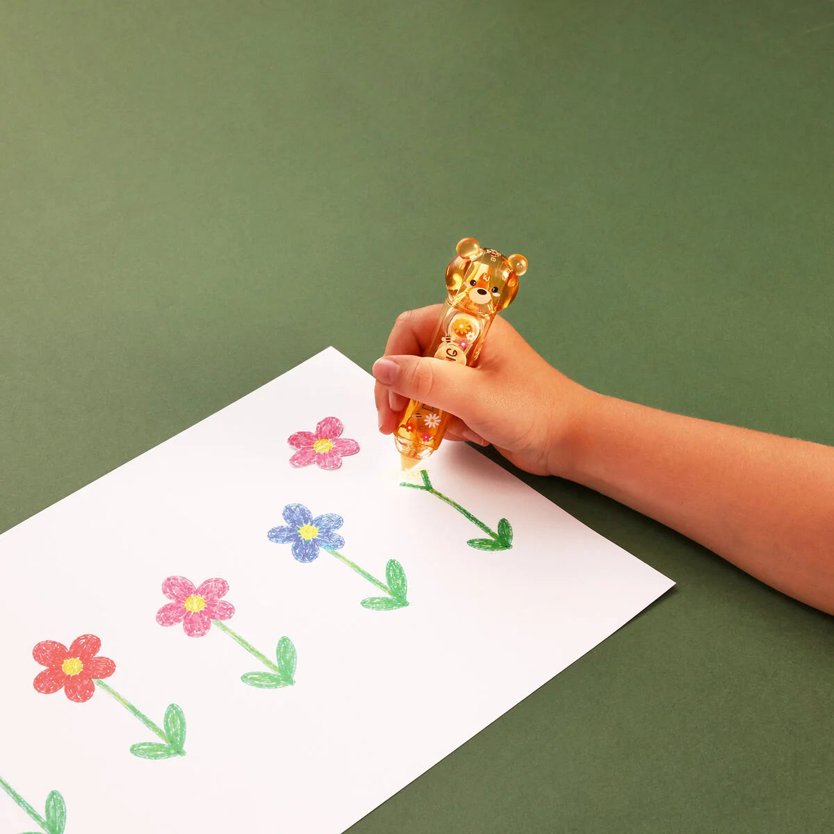 Hand hält den Zwei-in-Eins Korrektur- und Kleberoller Bear Sweet Bear von Legami über einem Blatt mit bunten gemalten Blumen vor grünem Hintergrund