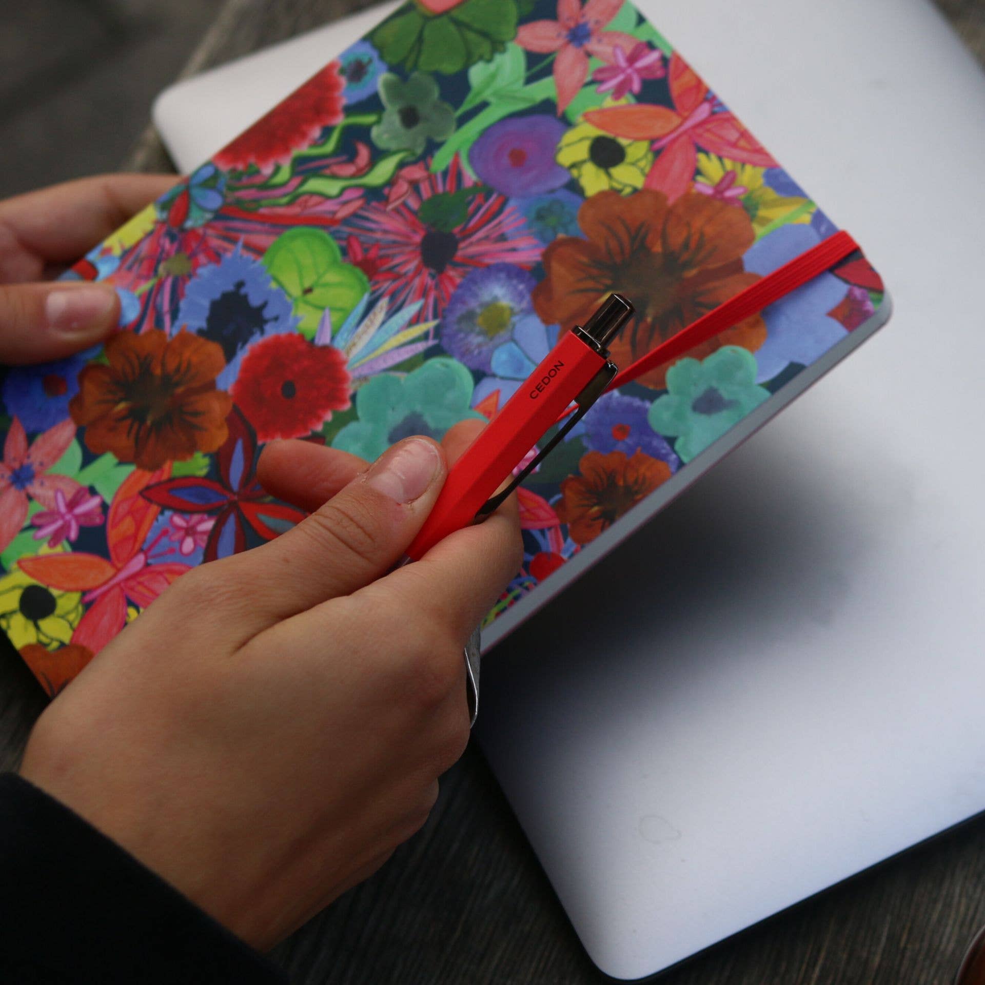 Cedon Kugelschreiber in Rot matt – in der Hand gehalten vor einem bunten Notizbuch mit Blumenmotiv