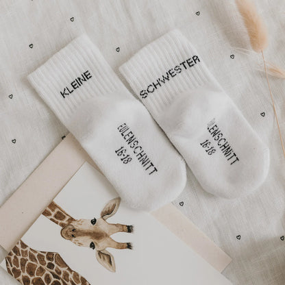 Eulenschnitt Baby-Socken „Kleine Schwester" in Weiß, Größe 16-18, auf herzgemustertem Stoff mit Giraffen-Karte bei Burgfrollein
