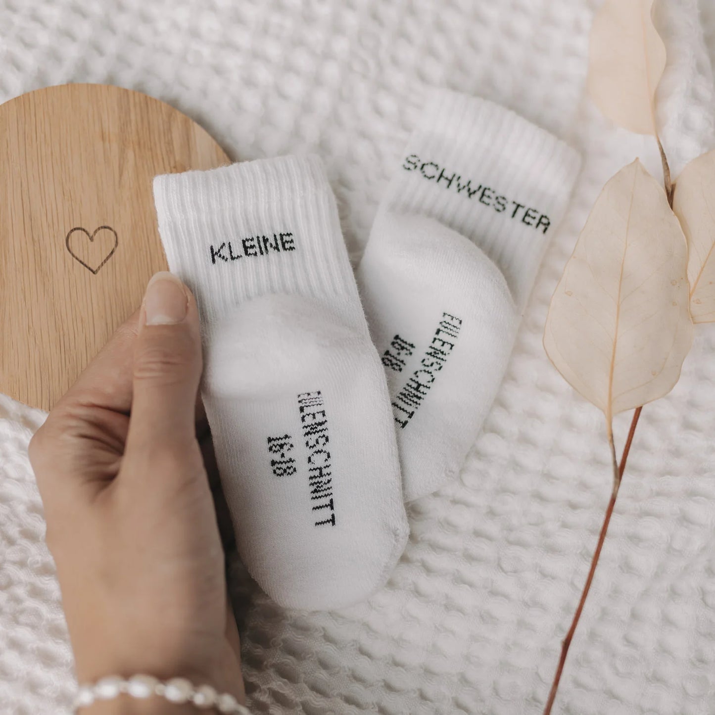 Hand hält Eulenschnitt Baby-Socken „Kleine Schwester" in Weiß, Größe 16-18, neben Holzschild mit Herz-Motiv bei Burgfrollein