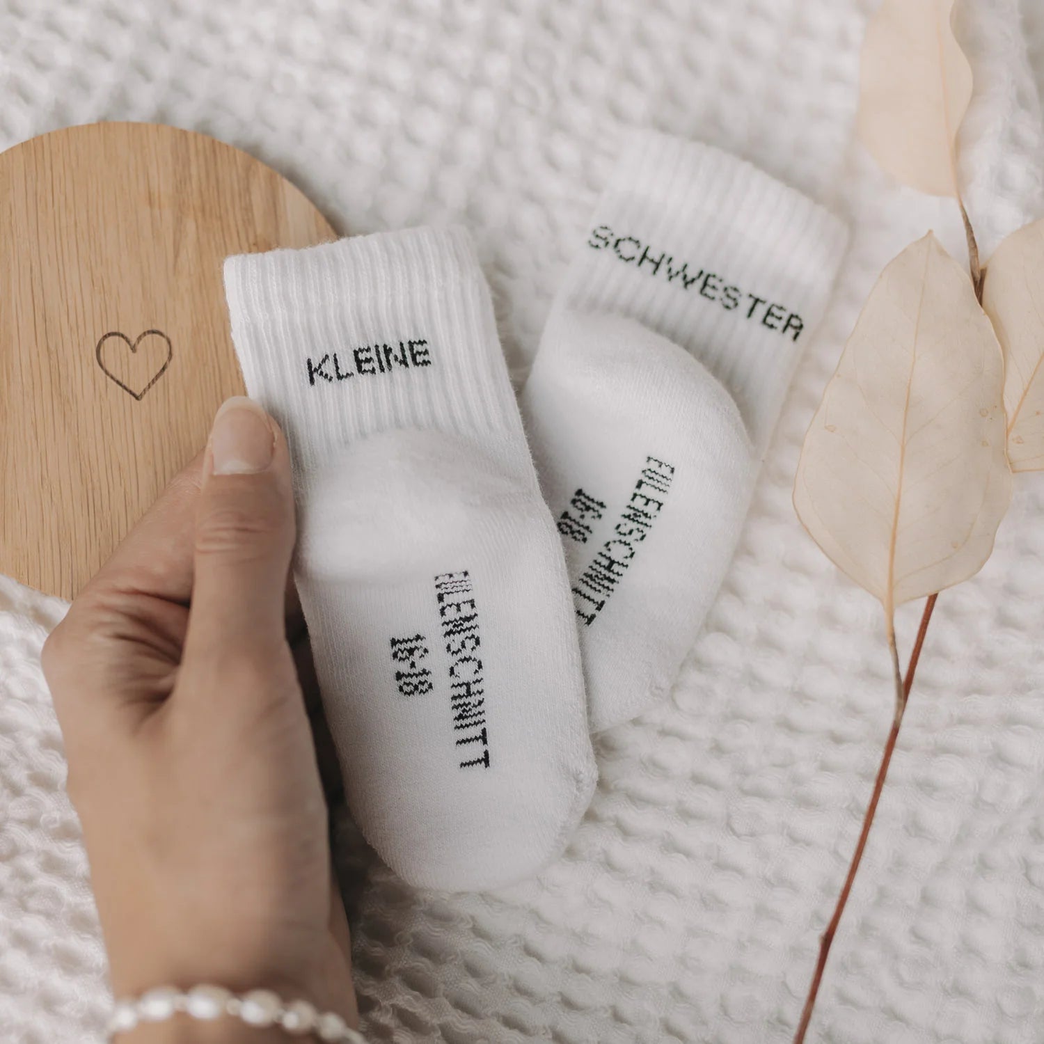 Hand hält Eulenschnitt Baby-Socken „Kleine Schwester" in Weiß, Größe 16-18, neben Holzschild mit Herz-Motiv bei Burgfrollein