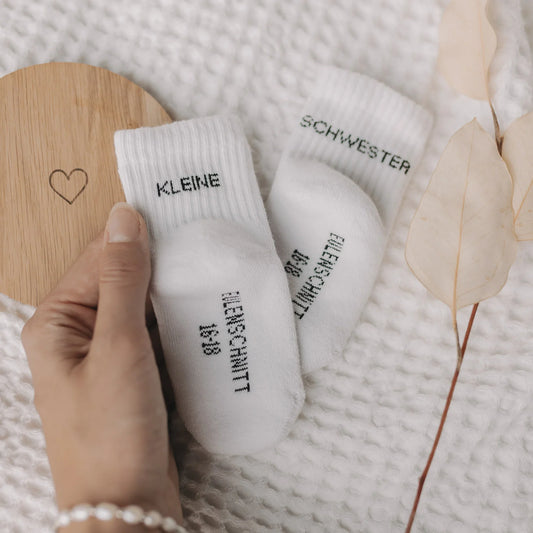 Hand hält Eulenschnitt Baby-Socken „Kleine Schwester" in Weiß, Größe 16-18, neben Holzschild mit Herz-Motiv bei Burgfrollein