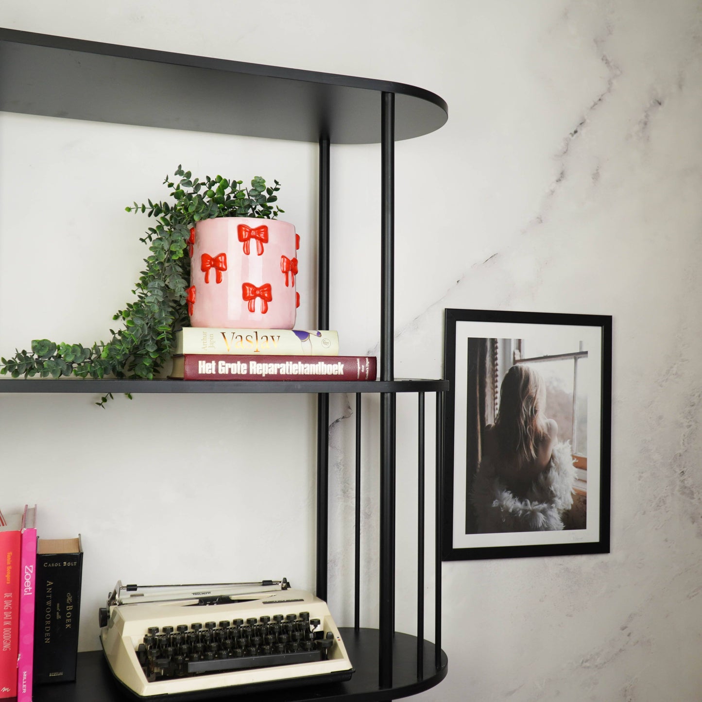 Rosa Blumentopf mit rotem Schleifen-Motiv von House Vitamin, bepflanzt mit einer Hängepflanze, dekoriert in einem schwarzen Regal vor Marmorwand mit Büchern, Vintage-Schreibmaschine und gerahmtem Foto – stilvoller Hingucker als Wohnungsdeko und Geschenkidee