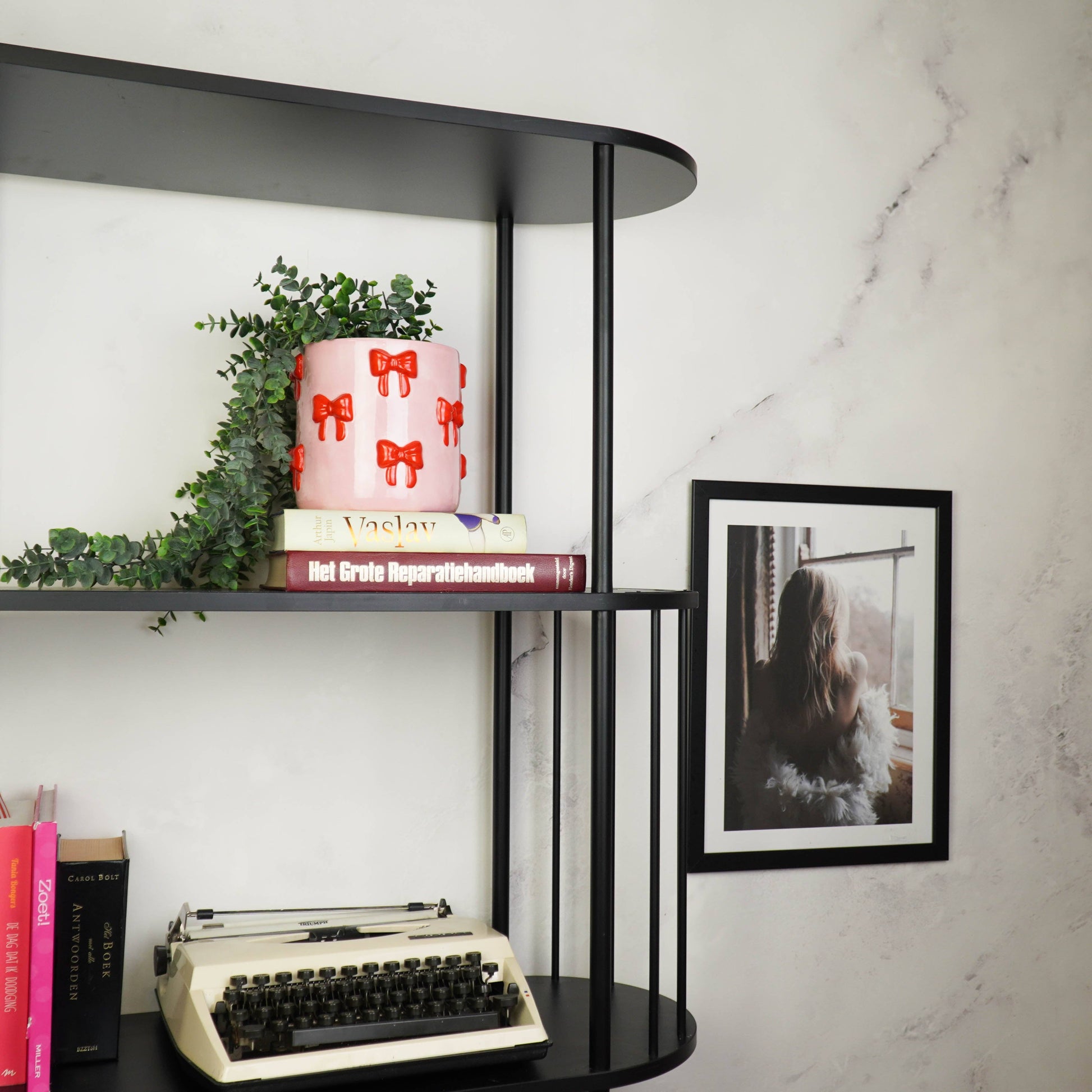 Rosa Blumentopf mit rotem Schleifen-Motiv von House Vitamin, bepflanzt mit einer Hängepflanze, dekoriert in einem schwarzen Regal vor Marmorwand mit Büchern, Vintage-Schreibmaschine und gerahmtem Foto – stilvoller Hingucker als Wohnungsdeko und Geschenkidee