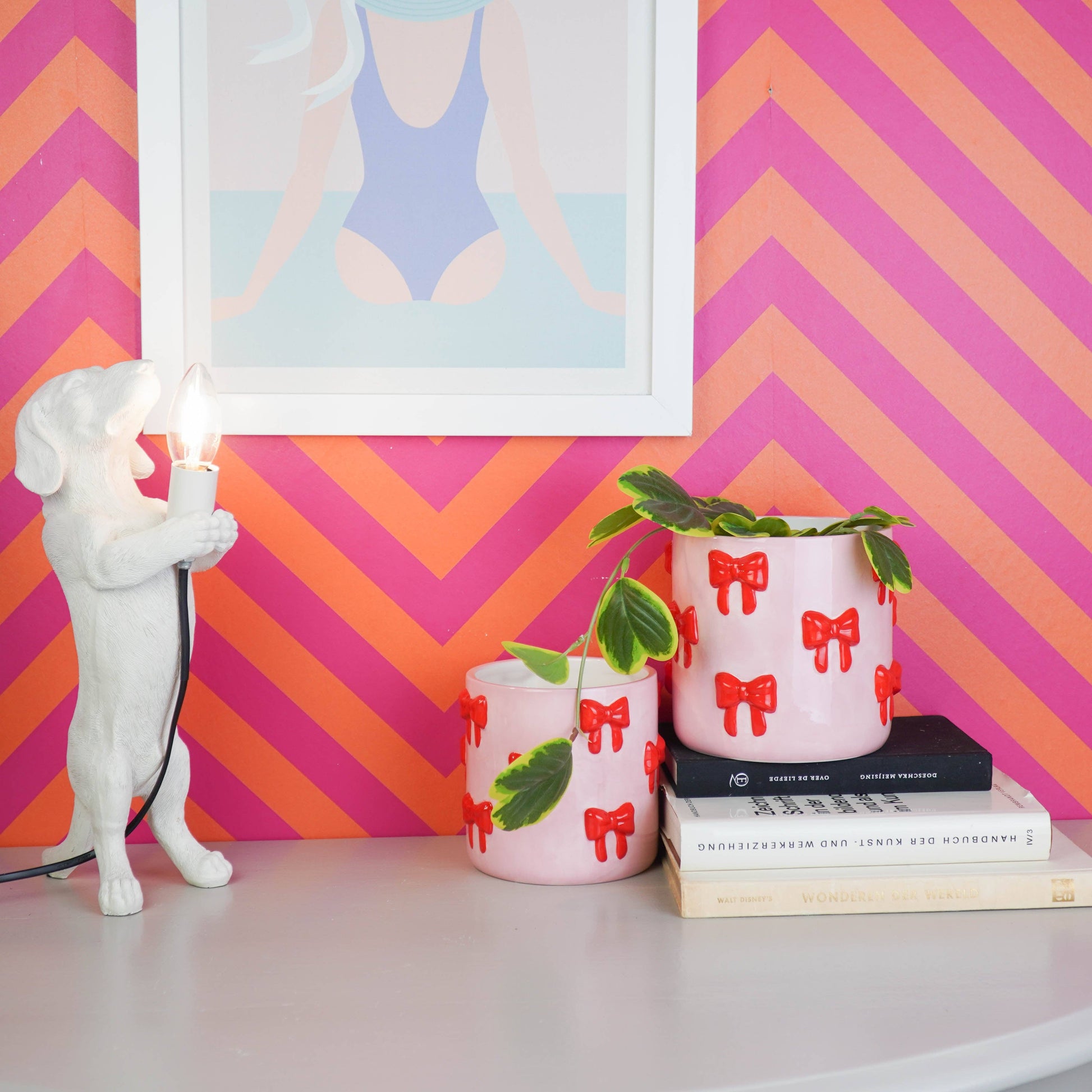 Zwei rosa Blumentöpfe mit rotem Schleifen-Motiv von House Vitamin, bepflanzt mit Grünpflanzen, in Szene gesetzt neben einer weißen Hund-Tischlampe vor pink-orangem Zickzack-Hintergrund – verspielter Hingucker als Wohnungsdeko und Geschenkidee