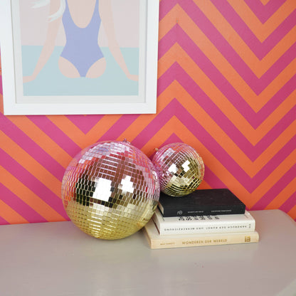 Discokugel „für gute Laune“ von House Vitamin (15 cm) in Rosé-Pink, daneben eine kleinere goldene Discokugel und ein Bücherstapel vor pink-orangem Zickzack-Hintergrund – funkelnder Hingucker als Wohnungsdeko und Geschenkidee