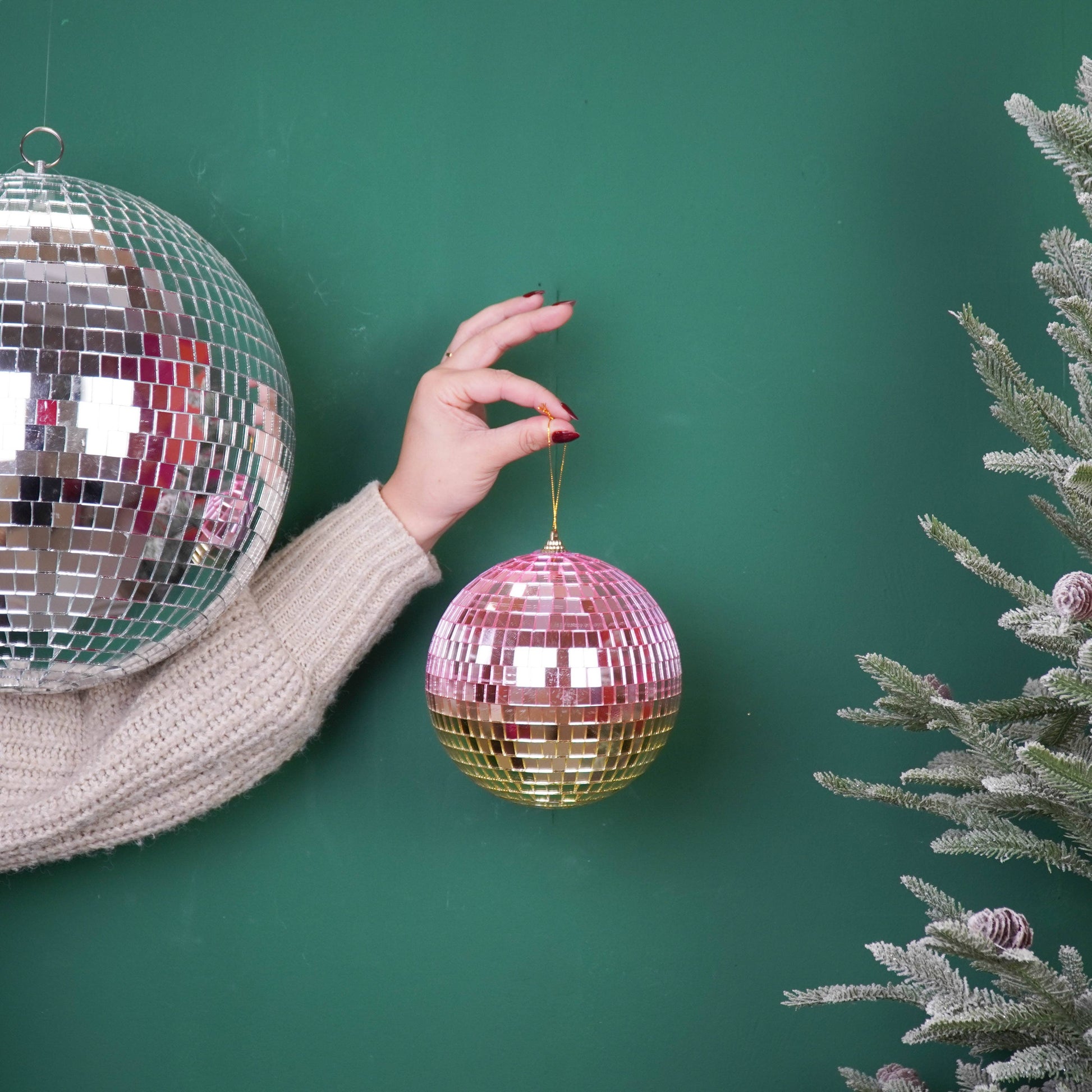 Discokugel „für gute Laune“ von House Vitamin (15 cm) in Rosé-Pink und Gold, an einer Schnur gehalten vor grünem Hintergrund mit großer silberner Discokugel und bereiftem Tannenzweig – funkelnder Hingucker als Weihnachtsdeko und Geschenkidee