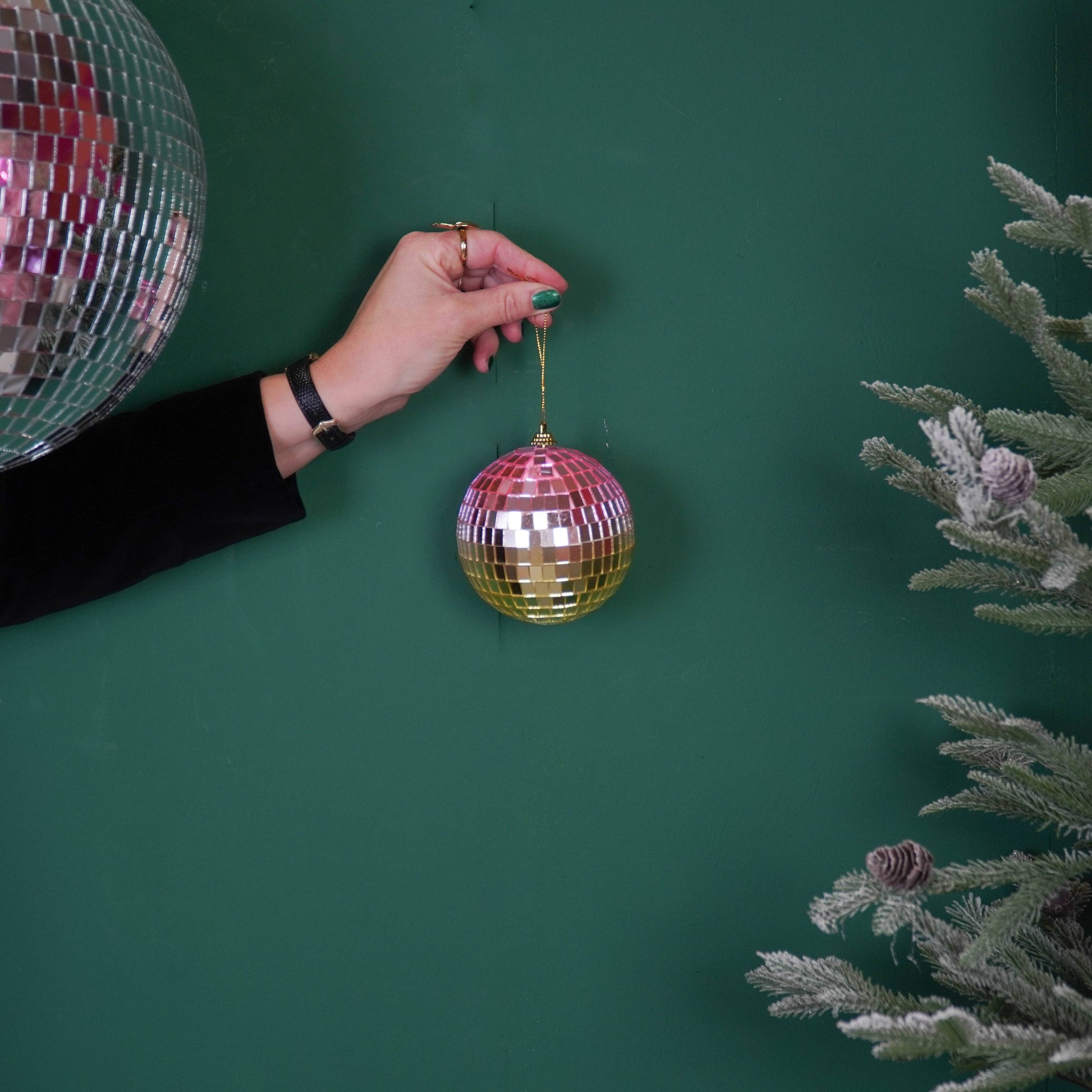 Discokugel für gute Laune von House Vitamin, 10 cm, in bunten Rosa-, Orange- und Goldtönen, an Schnur in der Hand gehalten neben silberner Discokugel und Tannenzweigen auf grünem Hintergrund – funkelnde Weihnachtsdeko und stimmungsvolle Geschenkidee