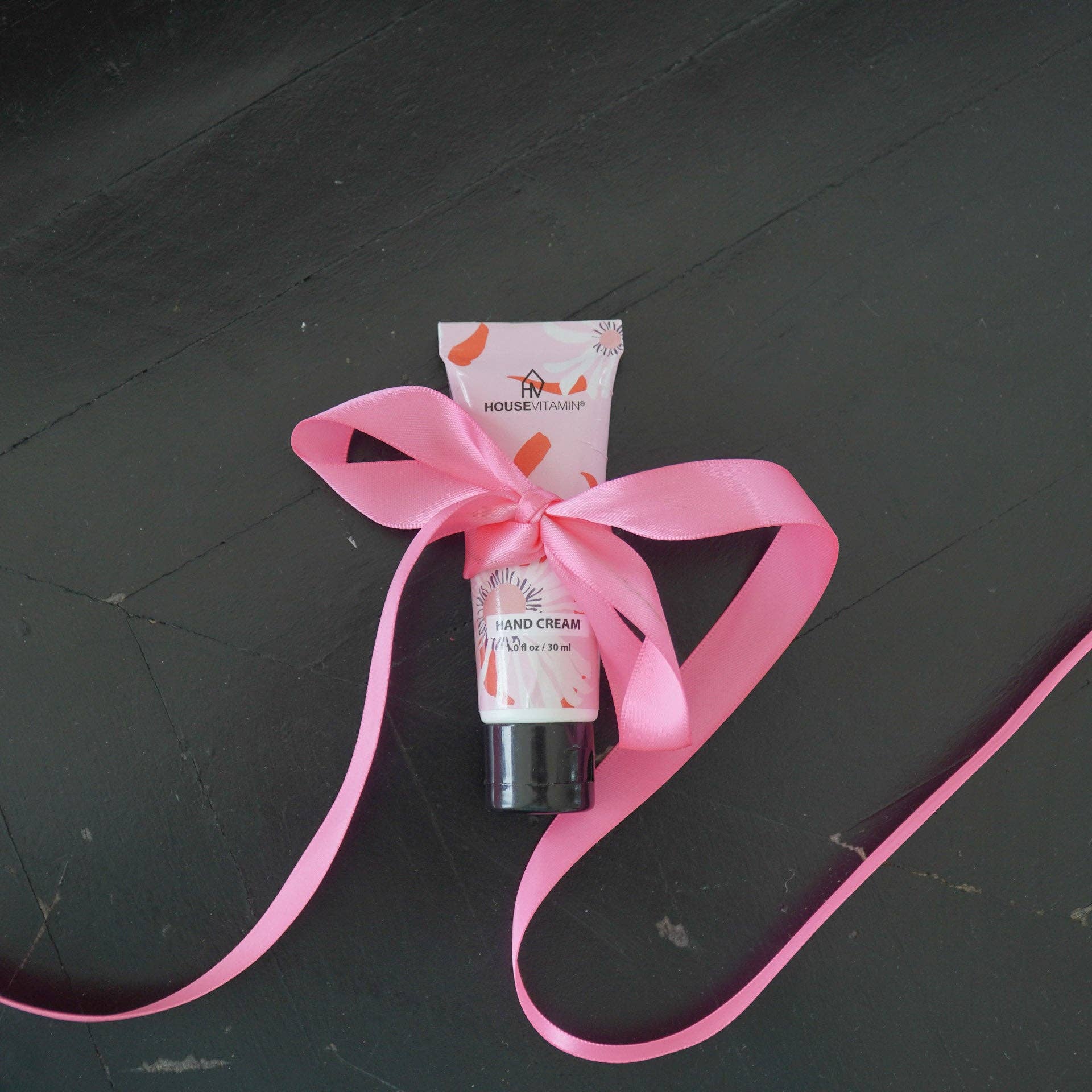 Kleine Tube Handcreme „Pfingstrosenblüte" von House Vitamin, 30 ml, mit großer gebundener rosa Schleife auf dunklem Holzuntergrund – florale Handpflege als festlich verpackte Geschenkidee