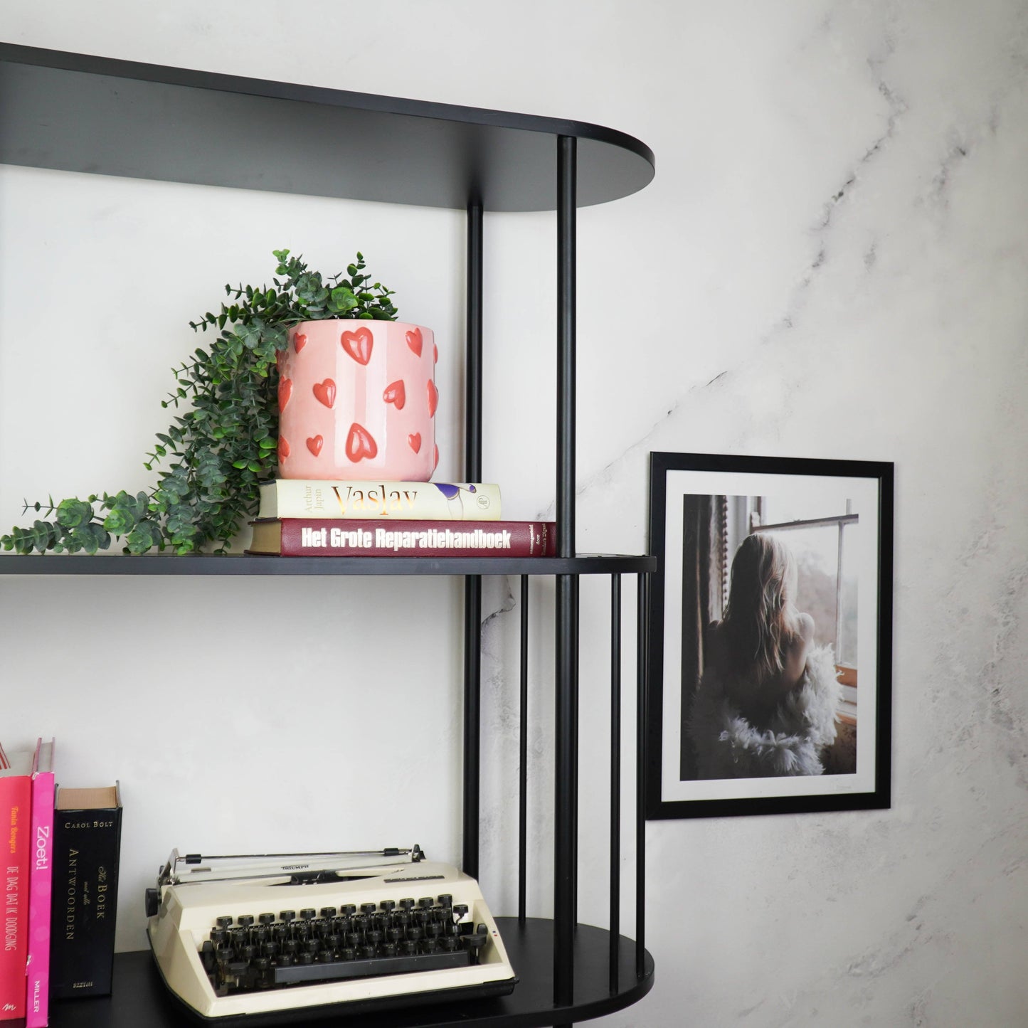 Rosa Keramik-Blumentopf mit rotem Herz-Motiv von House Vitamin, bepflanzt mit einer Hängepflanze, dekoriert in einem schwarzen Regal vor Marmorwand mit Büchern, Vintage-Schreibmaschine und gerahmtem Foto – romantischer Hingucker als Wohnungsdeko und Geschenkidee