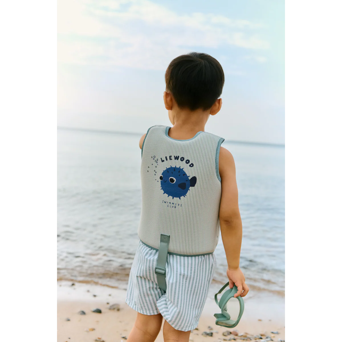 Kind am Strand von hinten mit Liewood Schwimmweste in Mint gestreift mit Kugelfisch-Print und Logo bei Burgfrollein