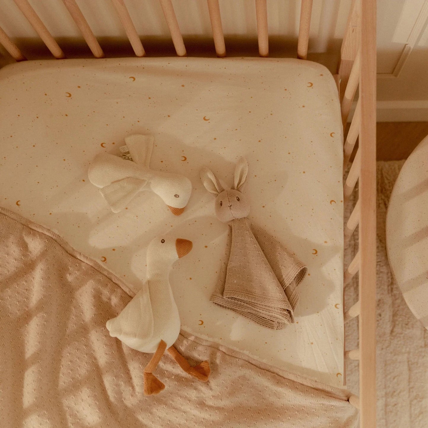 Little Dutch Geschenkset aus der Newborn Naturals Kollektion in Aktion – Gans-Kuscheltier, Gans-Rassel und Schmusetuch Hase liegen im Babybett auf einem Spannbettlaken mit Mond-Print