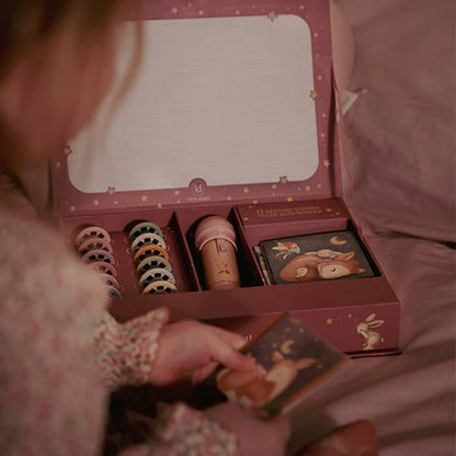 Mädchen sitzt vor der geöffneten rosa Geschenkbox des Little Dutch Geschichten-Taschenlampen-Sets aus der Fairy Garden Kollektion und betrachtet ein Geschichtenheft, daneben Diascheiben und Taschenlampe