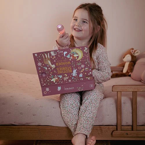 Mädchen sitzt im Bett und hält lächelnd die leuchtende Little Dutch Geschichten-Taschenlampe aus der Fairy Garden Kollektion in Rosa sowie die passende rosa Geschenkbox in den Händen