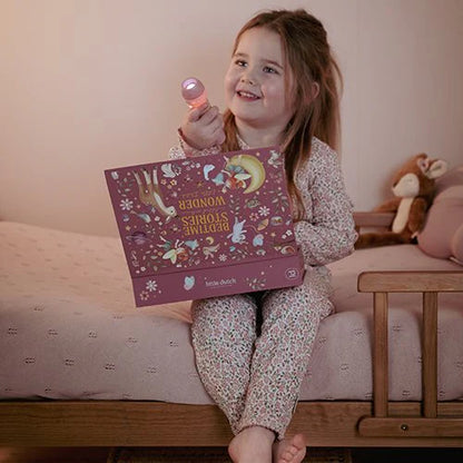 Mädchen sitzt im Bett und hält lächelnd die leuchtende Little Dutch Geschichten-Taschenlampe aus der Fairy Garden Kollektion in Rosa sowie die passende rosa Geschenkbox in den Händen