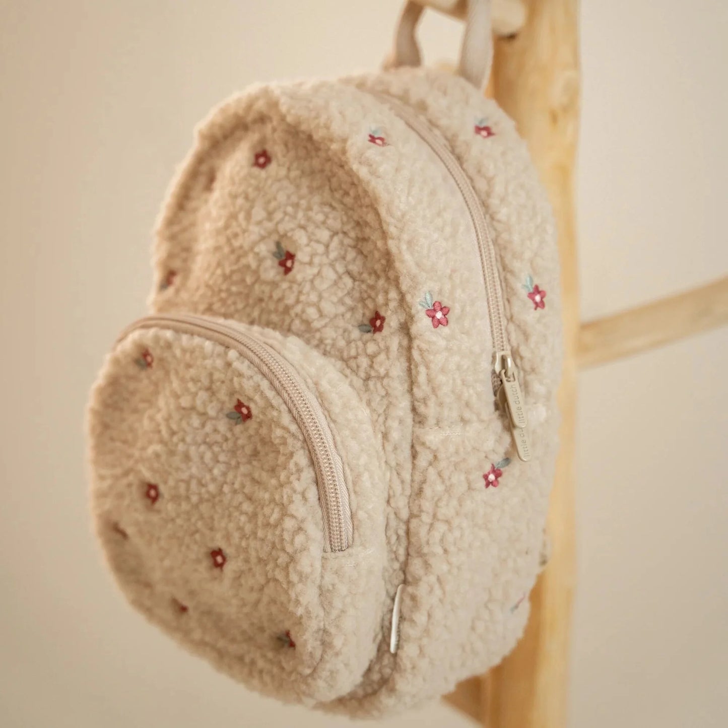 Little Dutch Teddy-Rucksack „Sand“ Detailansicht – Kinderrucksack aus weichem Teddyplüsch in Beige mit bestickten Blümchen und Reißverschluss, hängend an einer Holzleiter