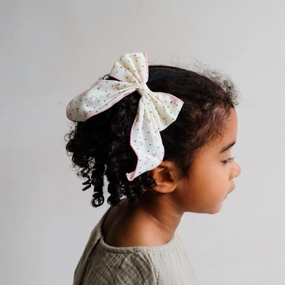 Haar-Schleife „Cherry" von Mimi & Lula, große weiße Stoffschleife mit Kirsch-Print und rosa Rand, getragen im Lockenhaar eines Kindes – kinderfreundlicher Haarschmuck und liebevolle Geschenkidee