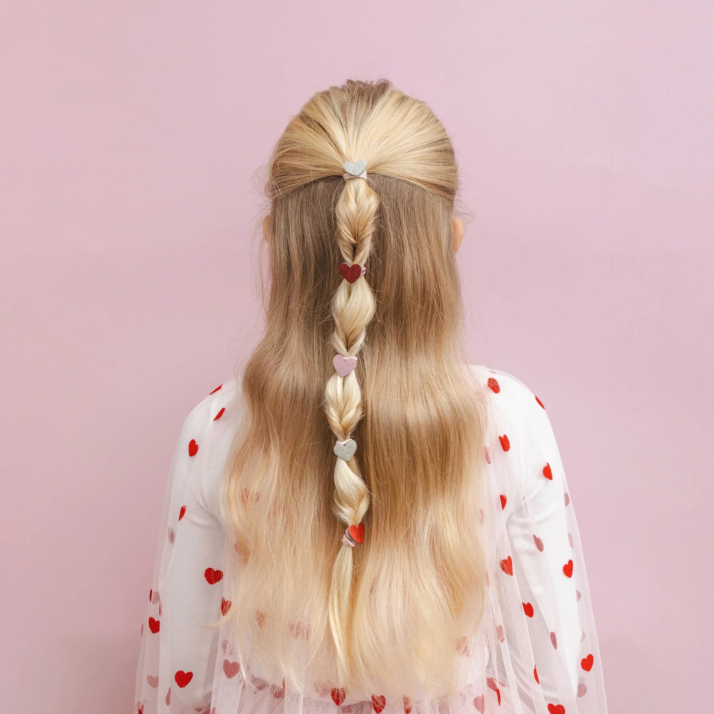 Kleine Haargummis mit Herz-Motiv von Mimi & Lula, 8er Set mit Stoff-Herzen in Gold-Glitzer, Rot, Rosa und Dunkelrot-Glitzer, getragen in einem geflochtenen Zopf im Haar eines Kindes mit Herz-Cape von hinten – romantischer Haarschmuck für Kinder und liebevolle Geschenkidee