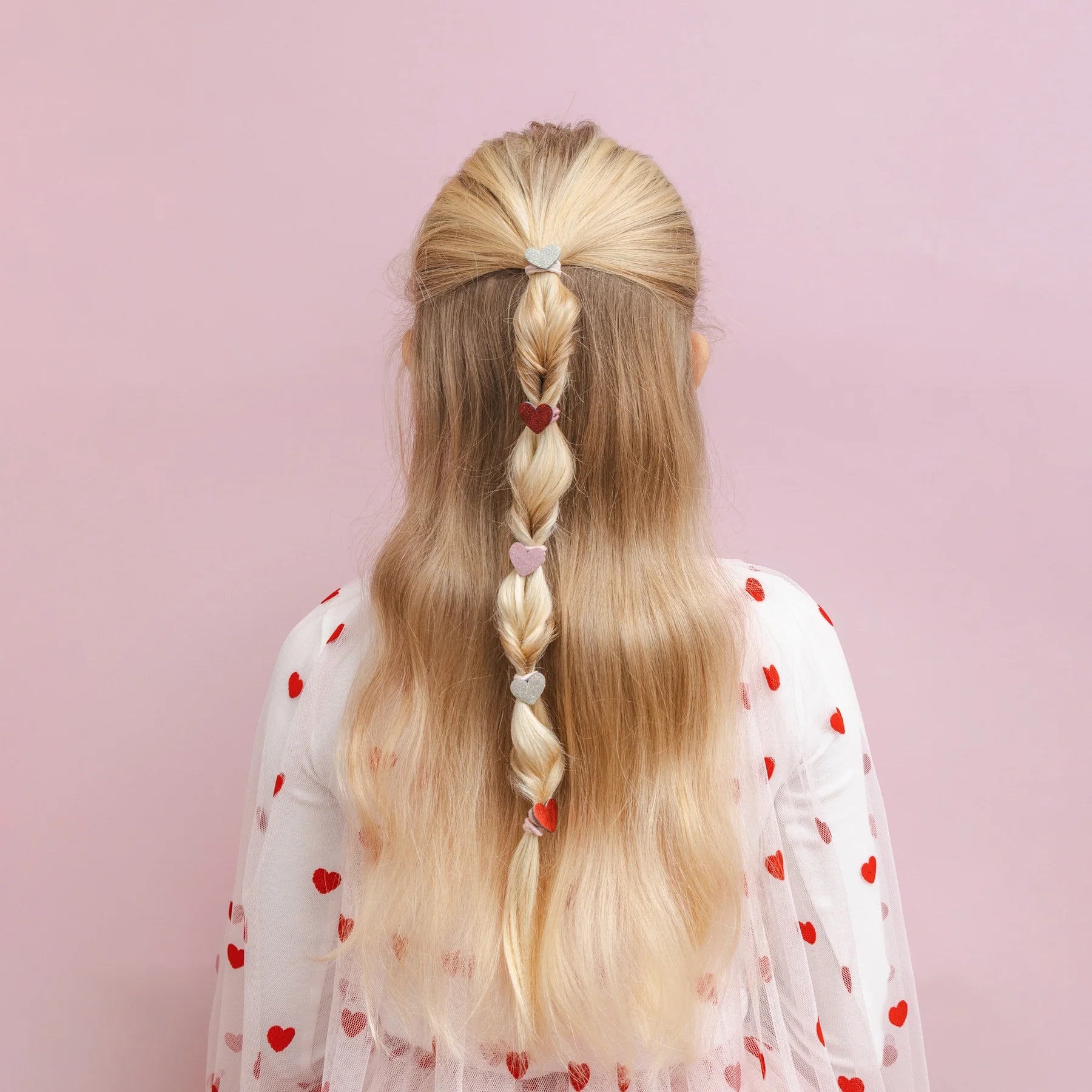 Kleine Haargummis mit Herz-Motiv von Mimi & Lula, 8er Set mit Stoff-Herzen in Gold-Glitzer, Rot, Rosa und Dunkelrot-Glitzer, getragen in einem geflochtenen Zopf im Haar eines Kindes mit Herz-Cape von hinten – romantischer Haarschmuck für Kinder und liebevolle Geschenkidee