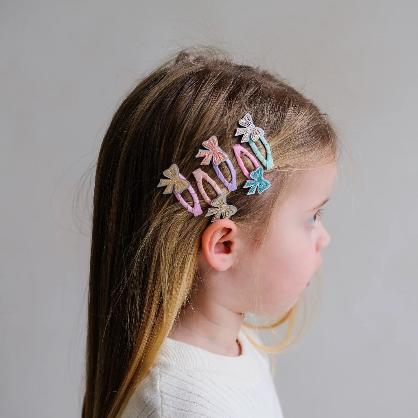 Glitzernde Haarspangen mit Schleifen-Motiv von Mimi & Lula, 5er Set mit Glitzer-Schleifen in Rosa, Gold, Pfirsich und Petrol auf Stoffspangen in Pastelltönen, nebeneinander getragen im Haar eines Kindes vor hellem Hintergrund – funkelnder Haarschmuck für Kinder und liebevolle Geschenkidee