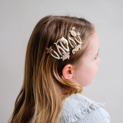 Haarspangen mit Meereswelt-Motiv von Mimi & Lula, 5er Set mit Krabbe, Delfin, Schildkröte, Seestern und Seepferdchen in Creme- und Goldtönen mit Glitzer-Details, getragen im Haar eines Kindes – kinderfreundlicher Haarschmuck und liebevolle Geschenkidee