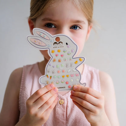 Ostern-Klebeohrringe von Mimi & Lula auf hasenförmiger Verpackungskarte mit zahlreichen Oster-Motiven wie Hasen, Eiern, Blumen und Schmetterlingen, gehalten von einem Kind – kinderfreundlicher Schmuck und liebevolle Geschenkidee zum Osterfest