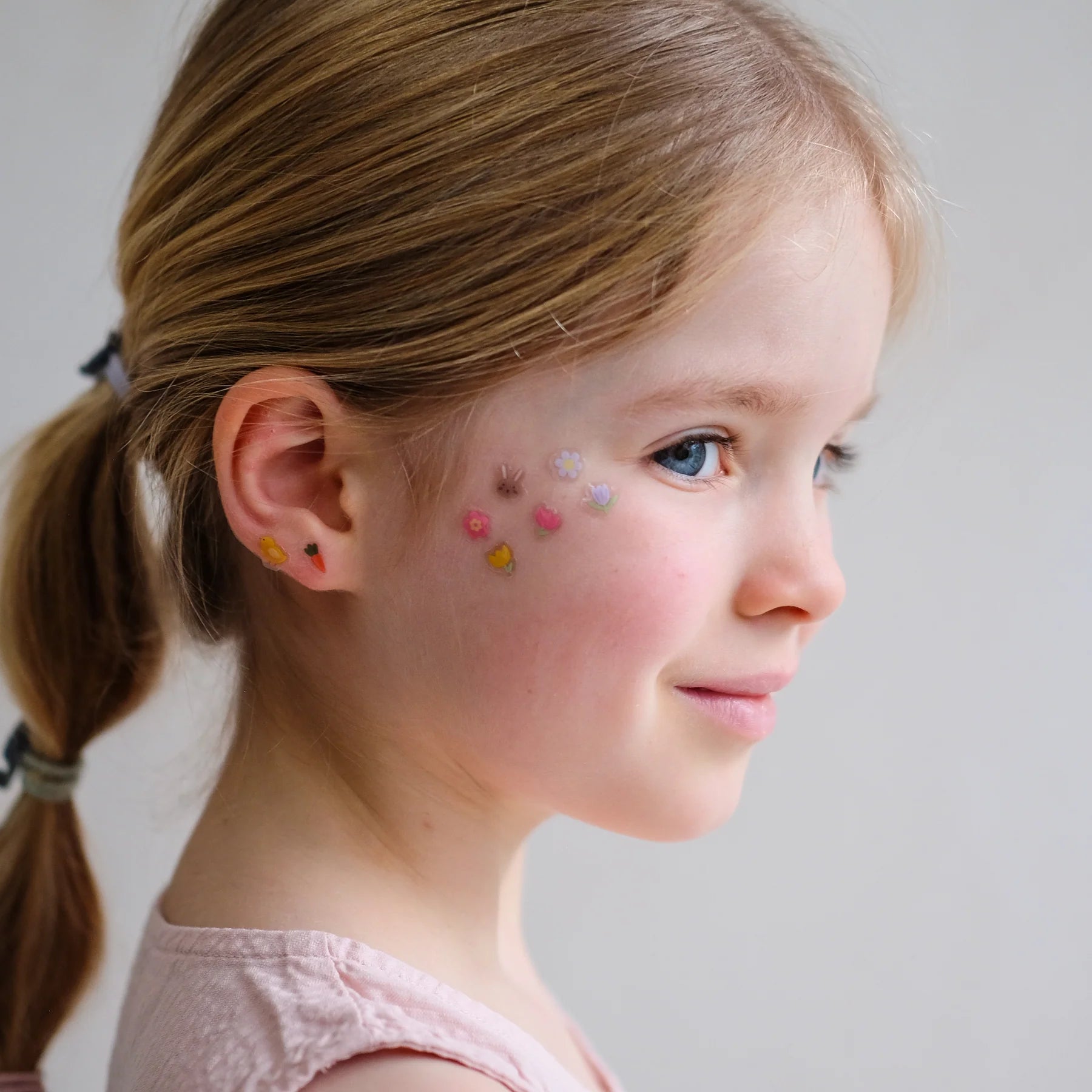 Ostern-Klebeohrringe von Mimi & Lula in bunten Pastelltönen, getragen am Ohr und als Mini-Sticker auf der Wange eines Kindes – kinderfreundlicher Schmuck mit Oster-Motiven und liebevolle Geschenkidee zum Osterfest