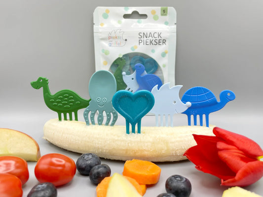 Snack Piekser Bouncy Five 5er Set von Pieksi und Griffel – Verpackung mit gemischten Pieksi-Figuren in Blau- und Grüntönen (Dino, Oktopus, Herz, Igel, Schildkröte) dekorativ auf einer Banane mit Obst und Tulpe arrangiert