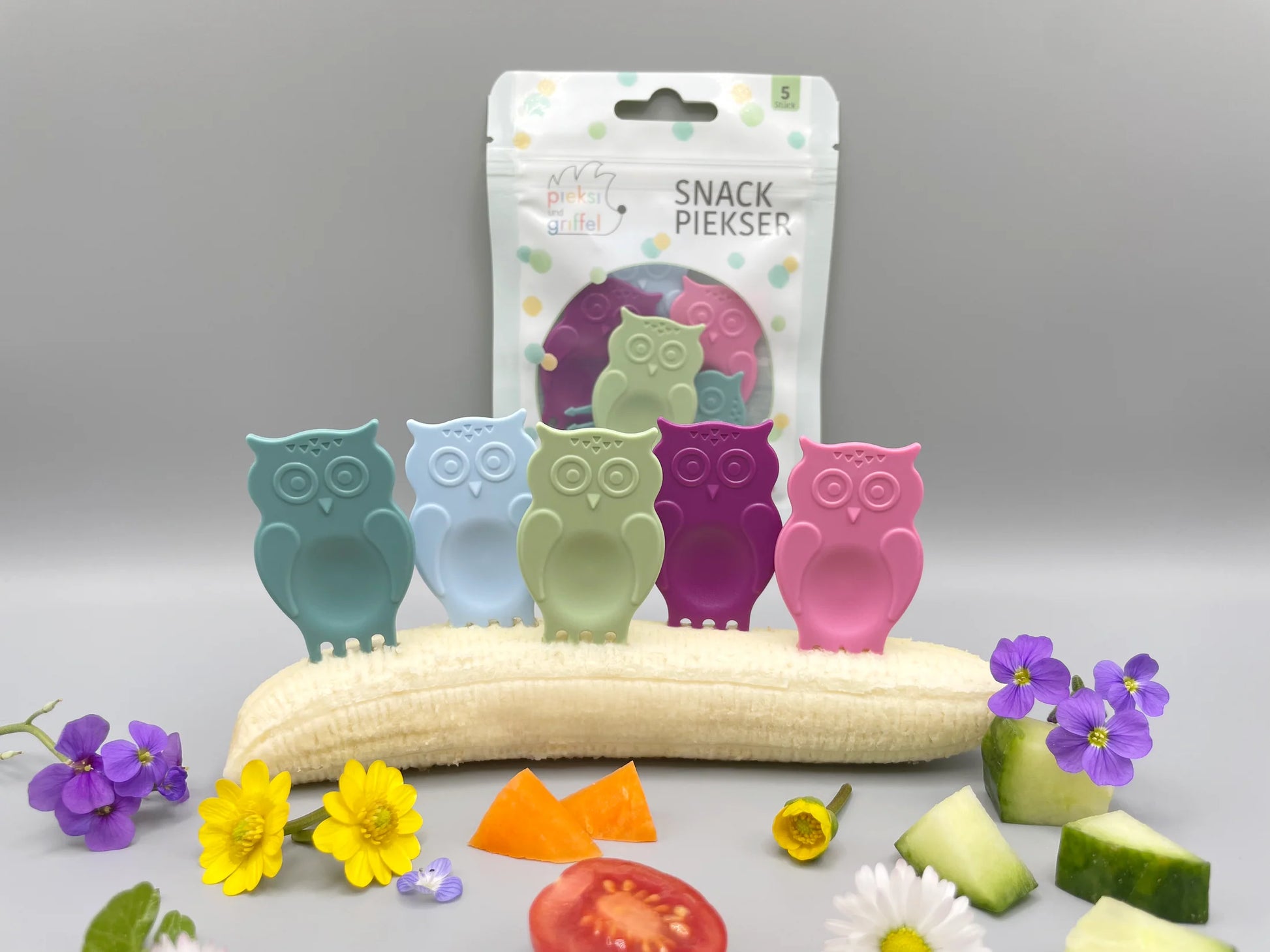 Snack Piekser Owl You Need 5er Set von Pieksi und Griffel – Verpackung mit den 5 Eulen-Pieksis in Pastellfarben dekorativ auf einer Banane mit Blüten, Tomate und Gurke arrangiert