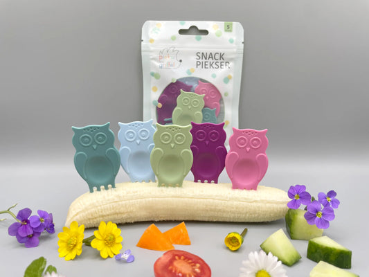 Snack Piekser Owl You Need 5er Set von Pieksi und Griffel – Verpackung mit den 5 Eulen-Pieksis in Pastellfarben dekorativ auf einer Banane mit Blüten, Tomate und Gurke arrangiert