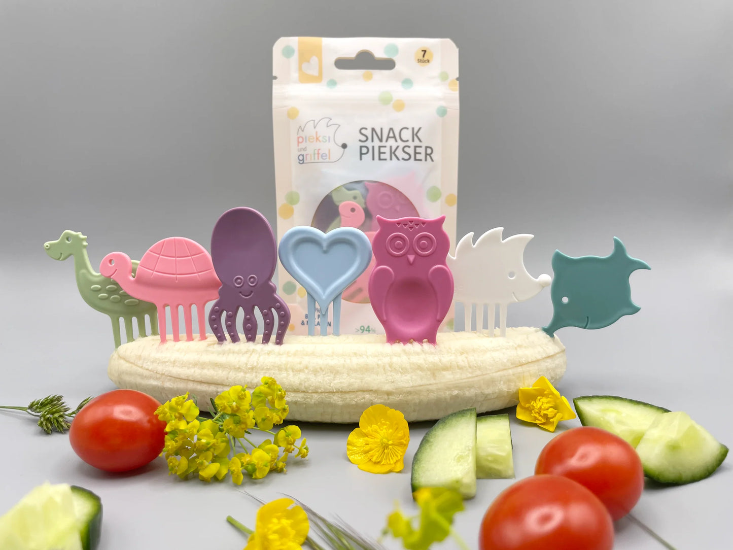 Snack Piekser Sweet & Soft 7er Set von Pieksi und Griffel – Verpackung mit den 7 Pieksi-Figuren in Pastellfarben dekorativ auf einer Banane mit Tomaten, Gurke und Blüten arrangiert