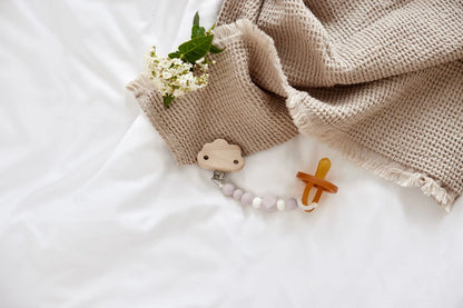 Rosa Schnullerkette „Little Universe“ von Lässig im Lifestyle-Setting – Holz-Silikon-Schnullerhalter mit Schnuller auf Babybettwäsche