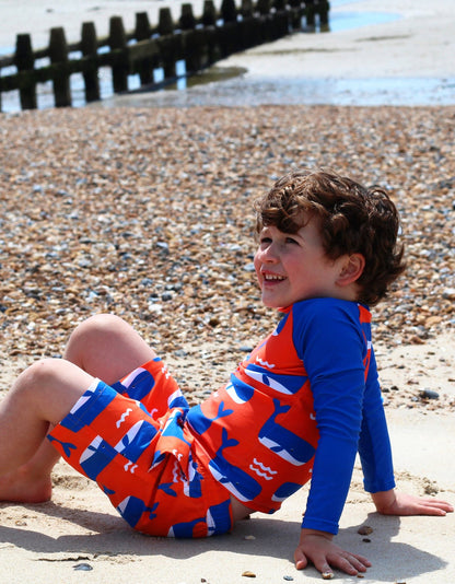Lachendes Kind am Strand im Toby Tiger UV-Schutz Shirt mit Wal- und Wellen-Print in Blau-Rot, UPF 50+ Badeshirt für Kinder